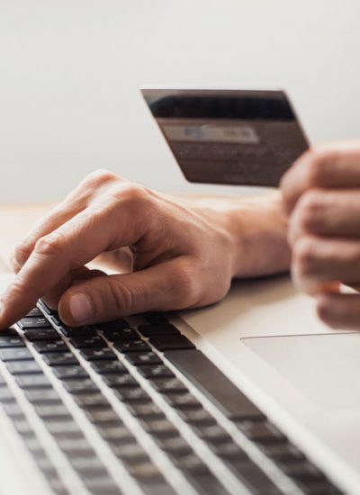 Person entering a key on the laptop keyboard while the other hand is holding a credit card. There are three packaged boxes on the laptop keyboard with shopping card icons.