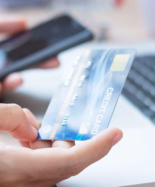 Person in front of a laptop computer holding a credit card on the right hand and a phone on the left hand.