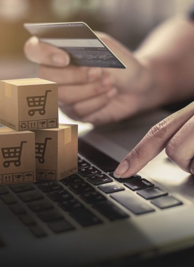 Person entering a key on the laptop keyboard while the other hand is holding a credit card. There are three packaged boxes on the laptop keyboard with shopping card icons.